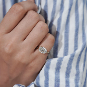 Close-up of a hand wearing an oval-cut diamond engagement ring in a solitaire setting, resting against a blue and white striped shirt. The diamond is secured with prongs and set on a slim yellow gold band.